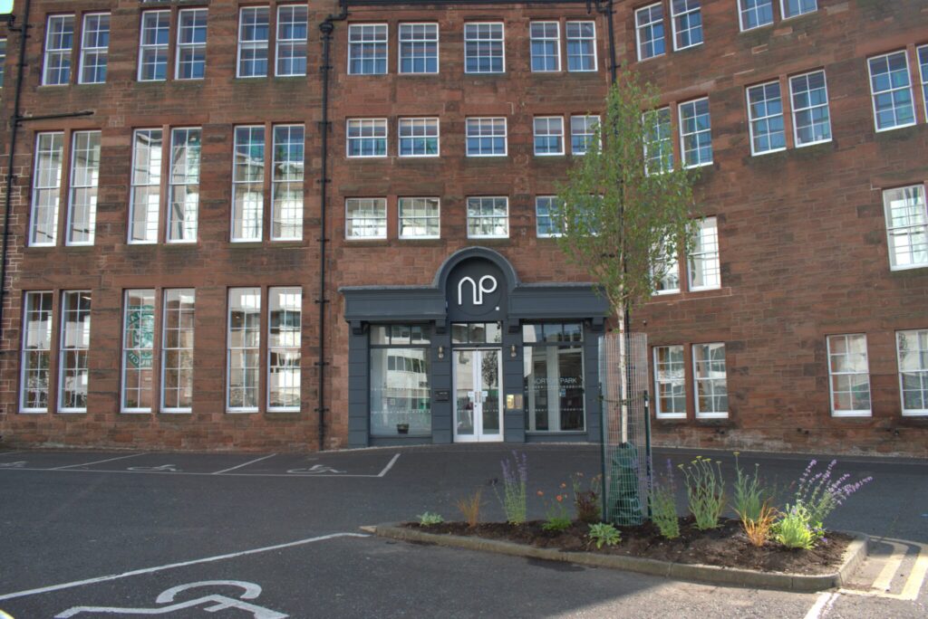 Front reception of Norton Park. Multistorey red sandstone building with grey entrance way. In the foreground, a triangle of spare space in the carpark now has a tree and low-level plants growing. 
