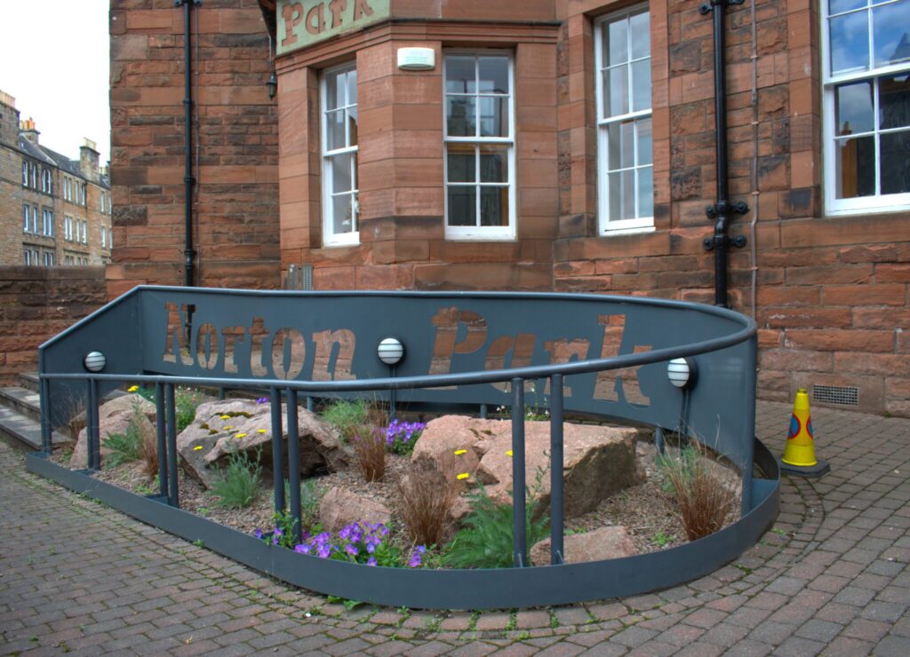 Red sandstone building in the background with paved path leading upwards. Grey railings include sign saying "Norton Park". Within the railings is a rockery with purple and yellow flowers growing.